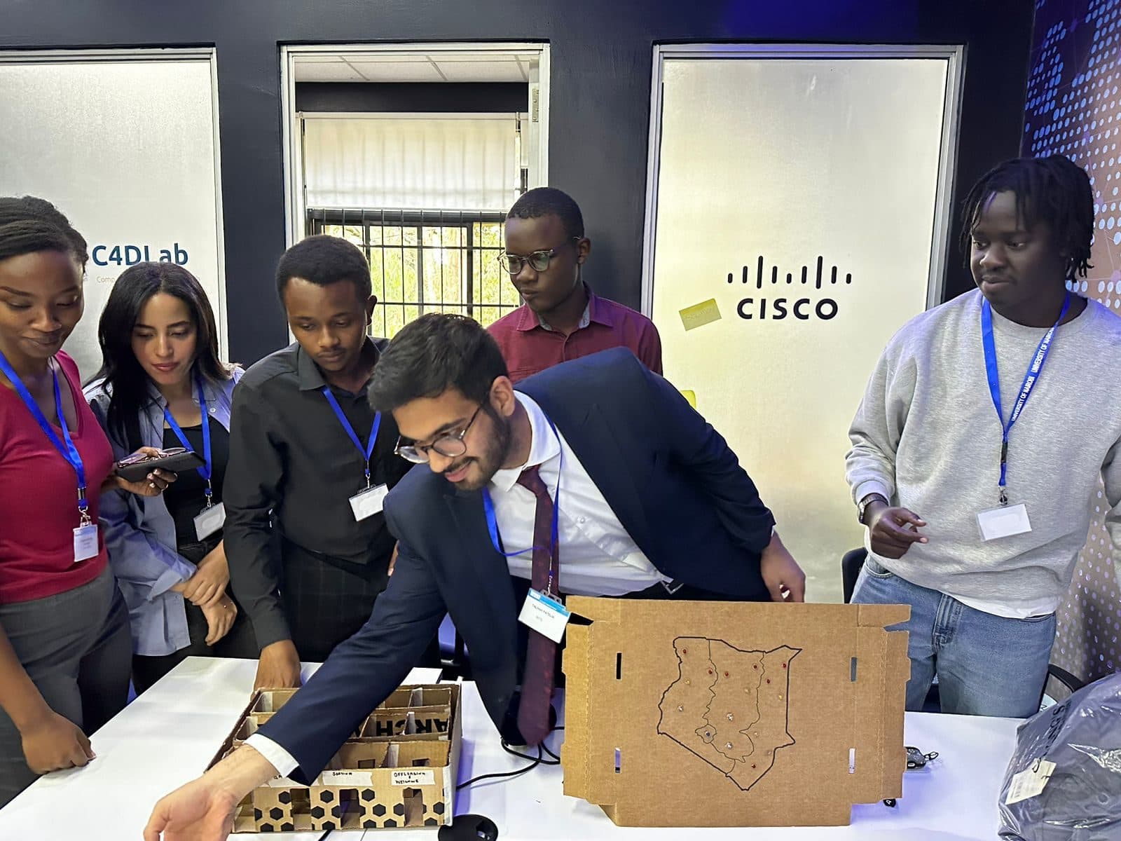 Talhah Patelia leaning over a cardboard model of the storage units while the team looks on, beside a cardboard map of Kenya