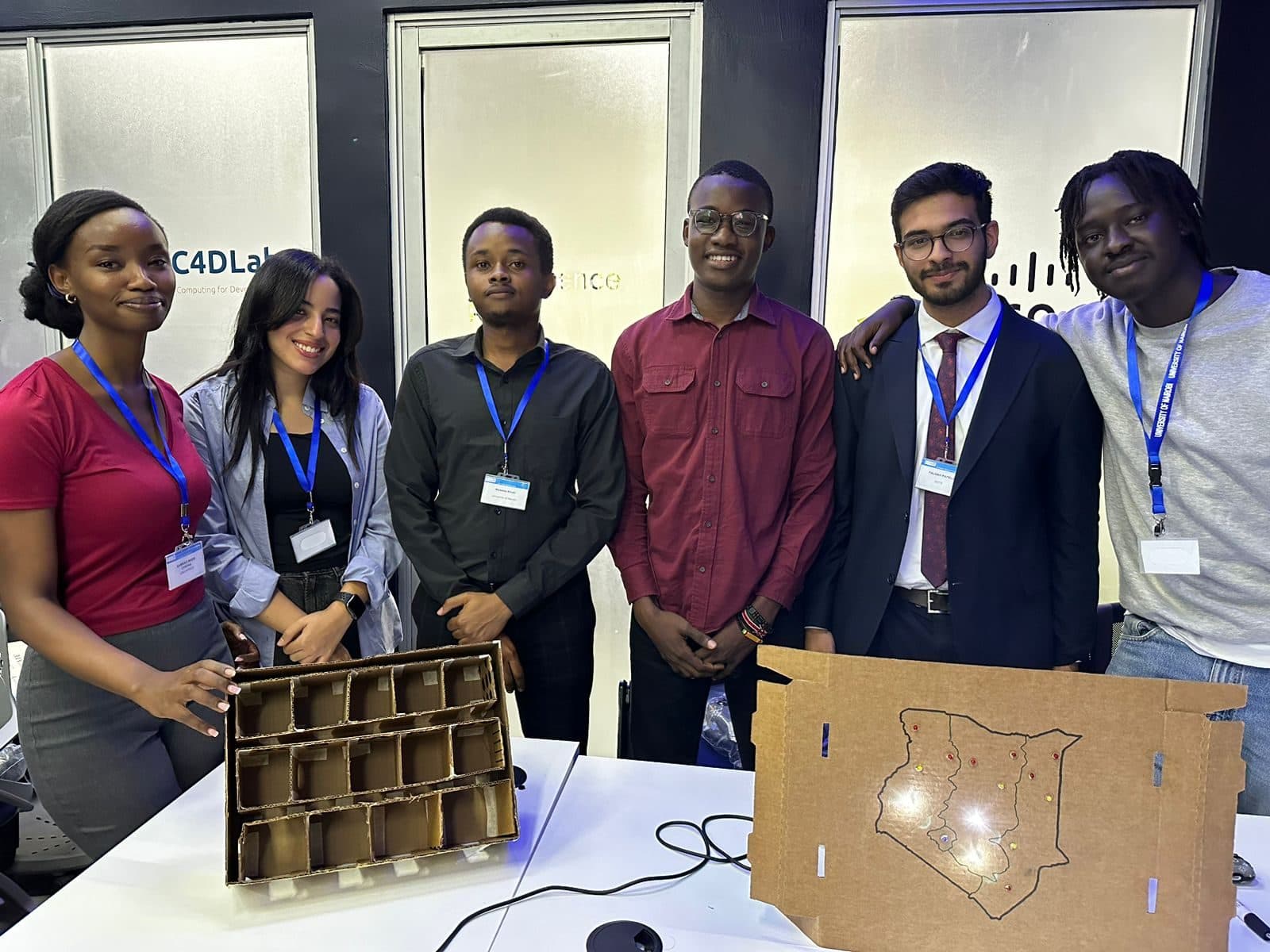 Talhah Patelia in a dark suit with five team members at the C4DLab in Nairobi, in front of a cardboard storage prototype and a cardboard map of Kenya