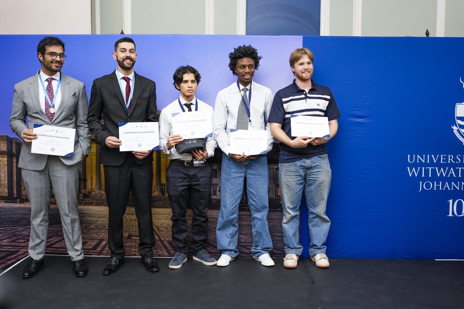 Talhah Patelia, Jared Swart and three other participants standing on stage holding certificates of recognition