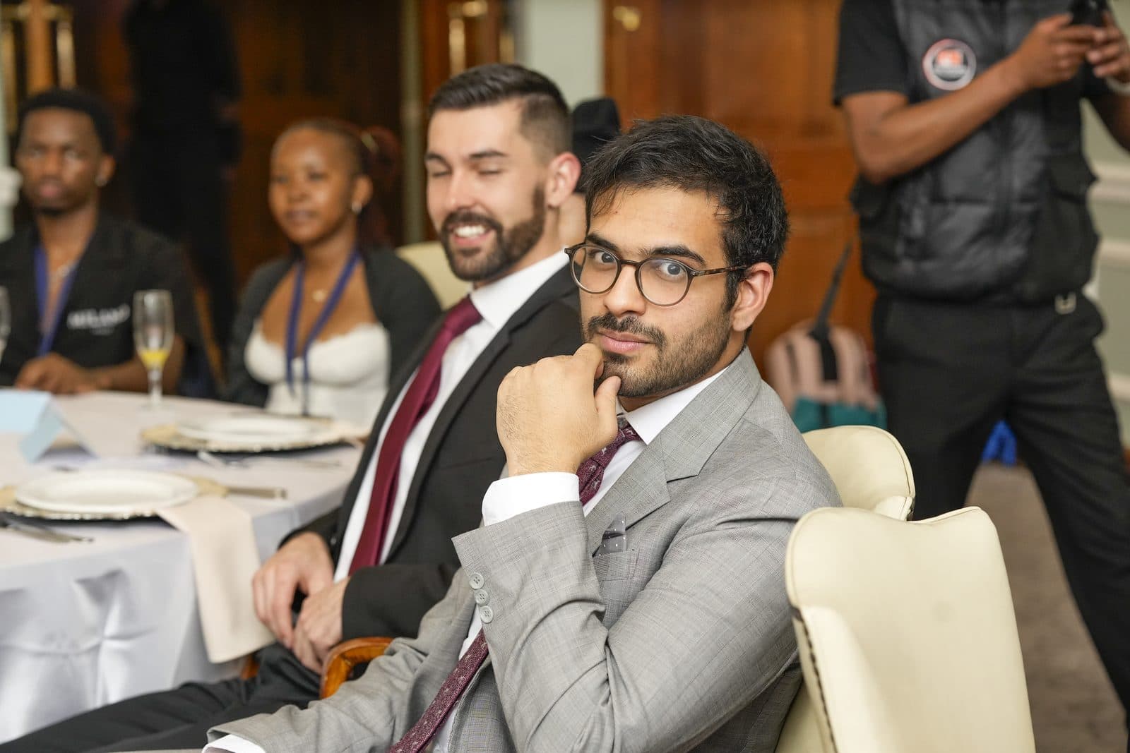 Talhah Patelia in a grey suit and round glasses, resting his chin on his hand, with Jared Swart smiling behind him at the AFRETEC SEED Challenge dinner
