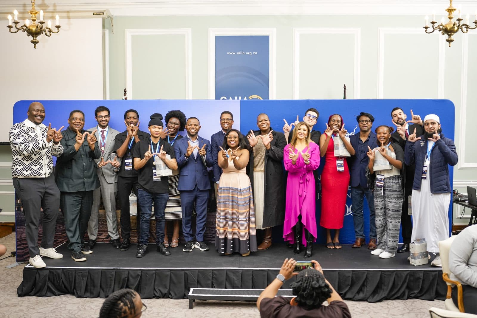 About twenty winners, coaches and guests on stage at Wits making W hand signs, with Talhah Patelia third from the left