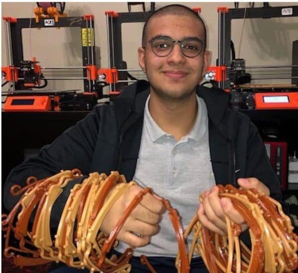 Talhah Patelia holding a stack of 3D-printed face shield frames in front of 3D printers
