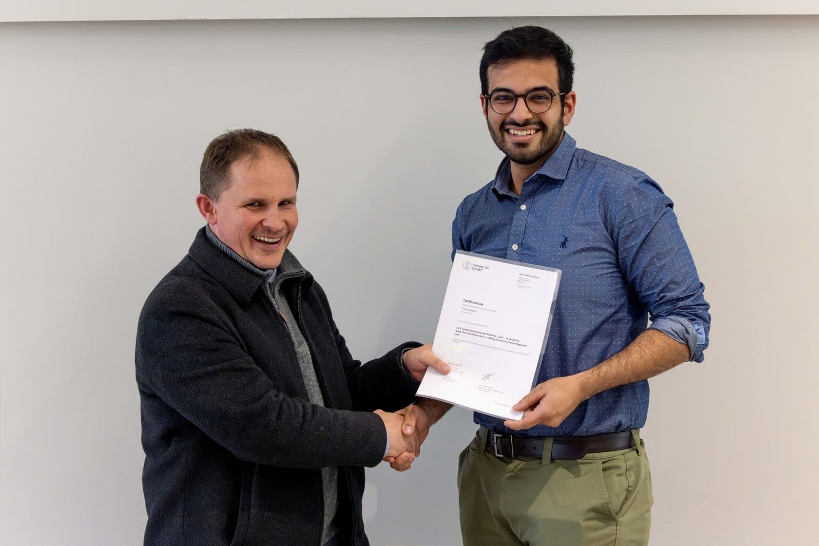 Talhah Patelia shaking hands with a man while holding his University of Zurich confirmation for the Deep Dive into Blockchain summer school