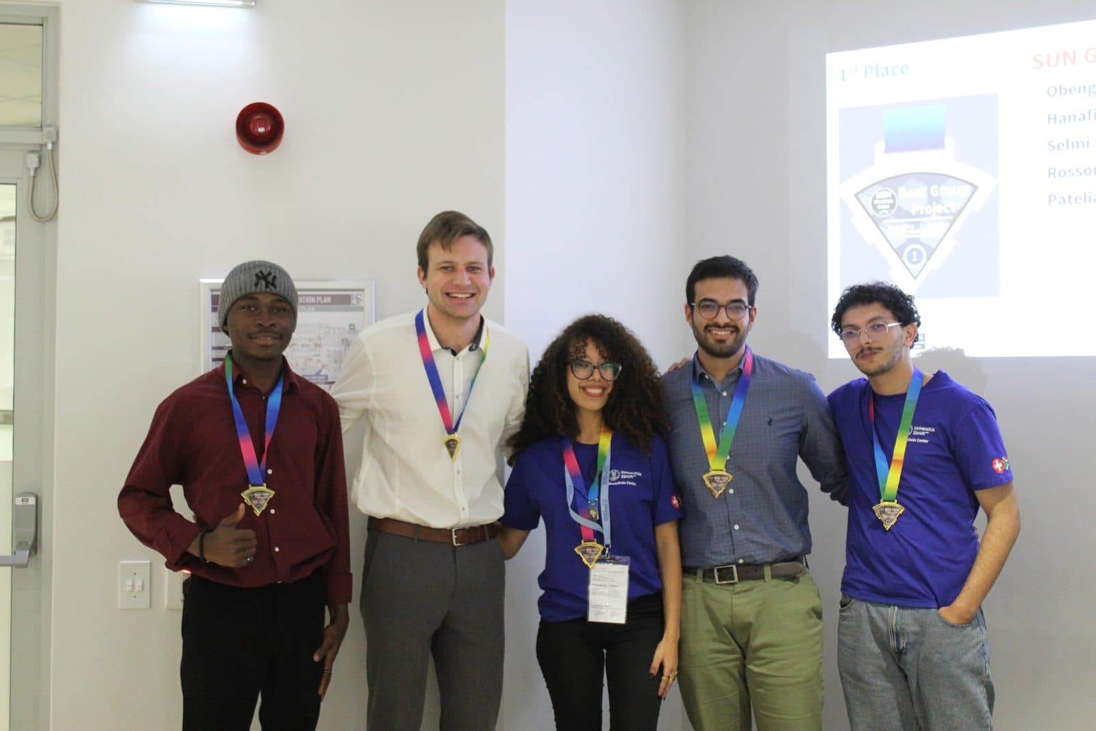 Five members of the winning team wearing gold medals on rainbow ribbons in front of a projected slide that lists them as first place in the best group project