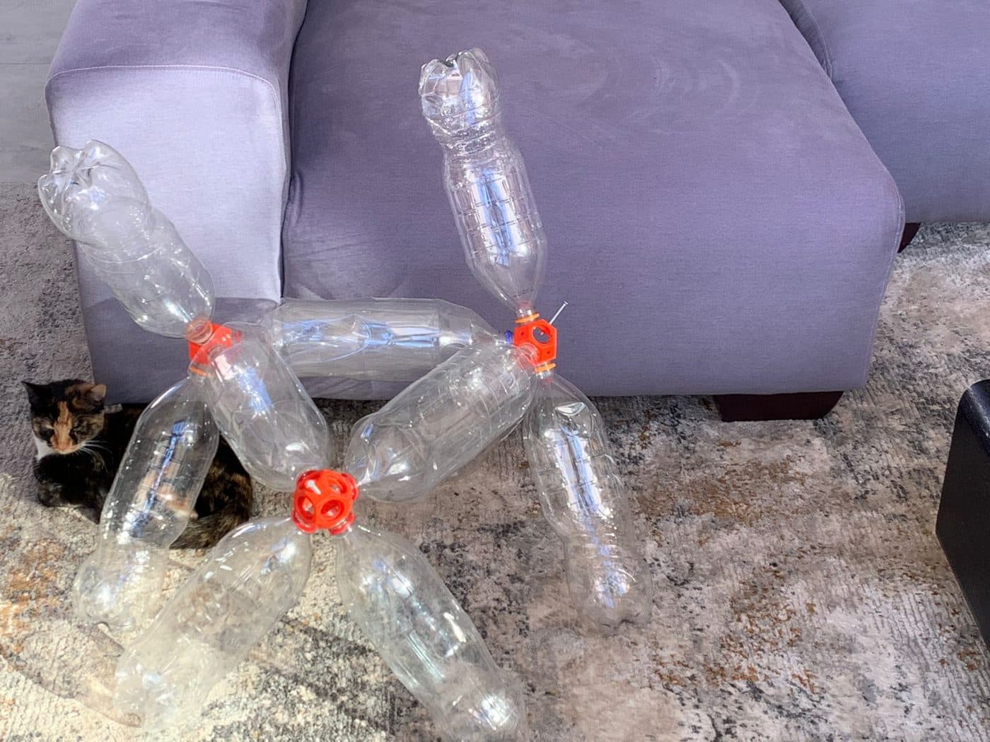 A small model made from plastic bottles and an orange connector, standing on a sofa